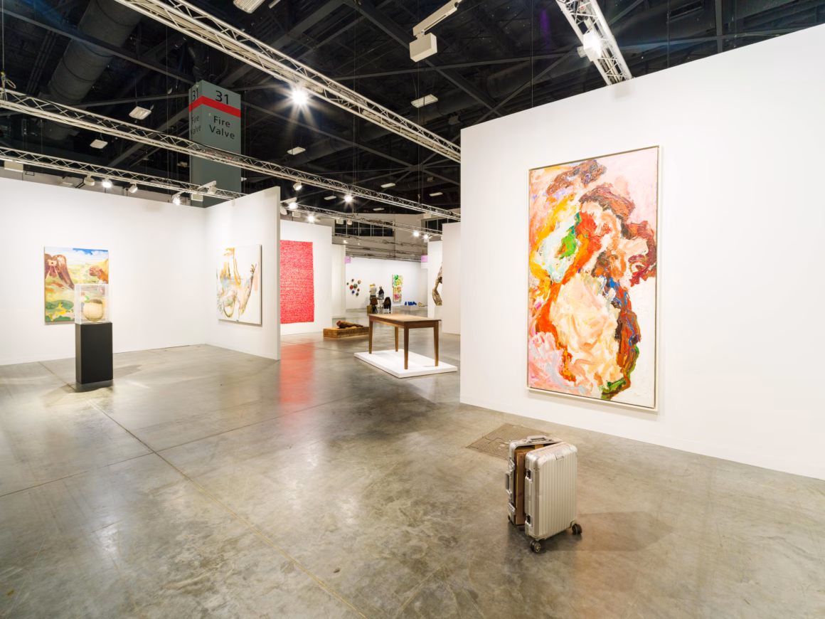 Contemporary art gallery booth at Art Basel Miami featuring colorful abstract paintings and sculptures with luxury silver suitcase in foreground