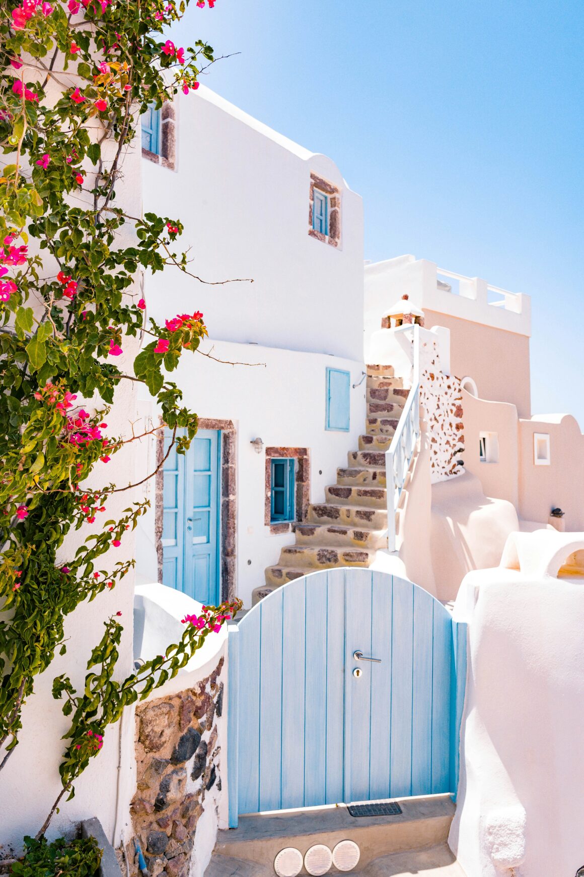 A bright, sunlit view of Oia, Greece, featuring whitewashed buildings, blue doors, stone steps, and pink bougainvillea—an ideal destination for slow travel and Mediterranean charm.