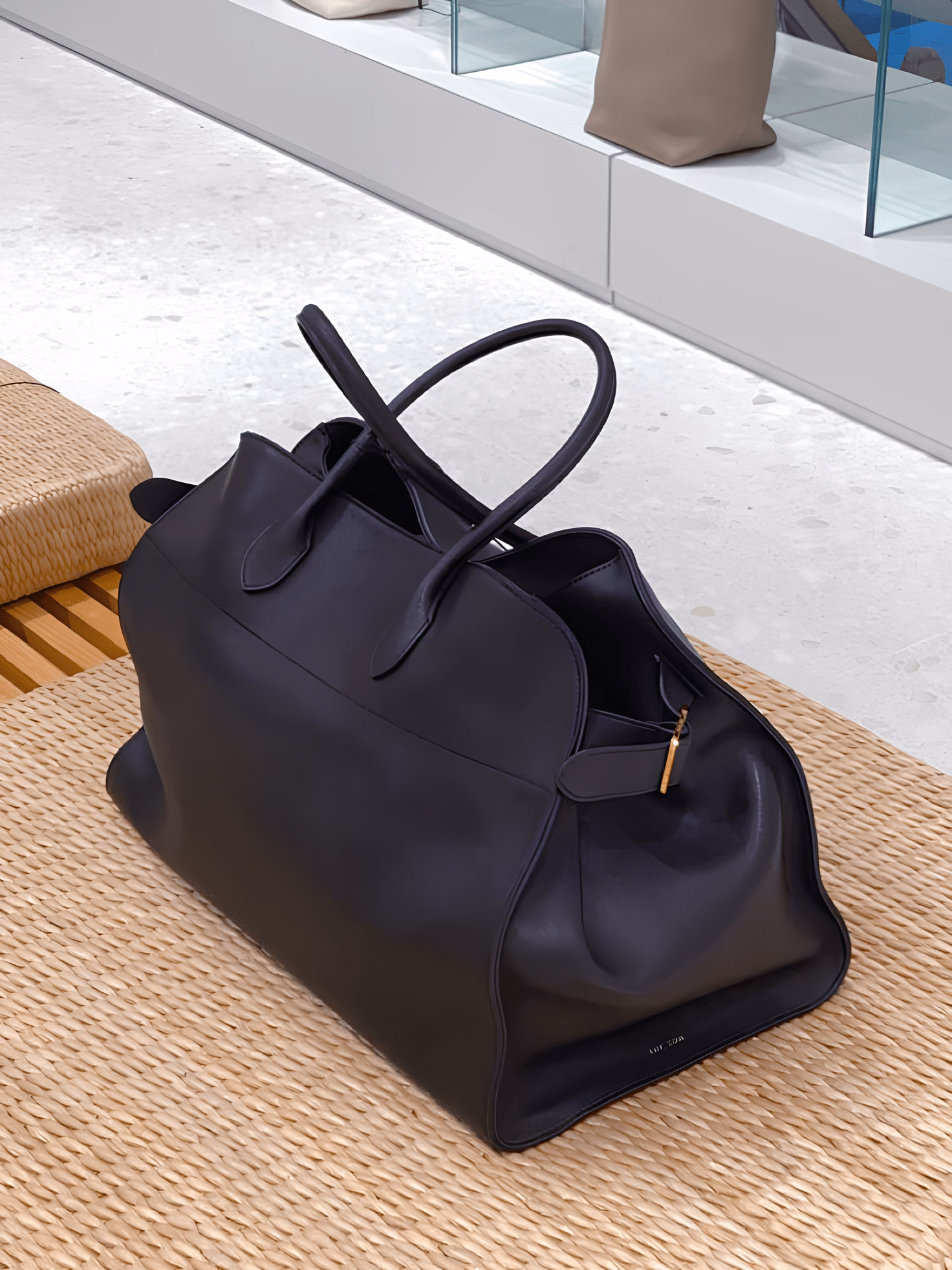 Soft-structured black leather Margaux handbag by The Row, shown resting on a woven bench in a neutral retail setting