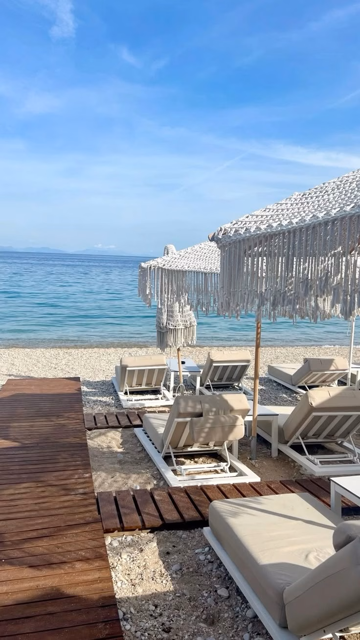 White lounge chairs and macramé umbrellas along a pebble beach with clear turquoise water at Verde Blu